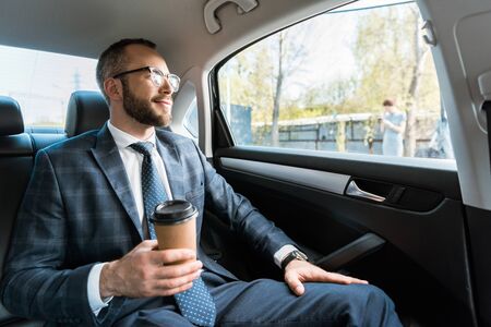 Happy businessman holding paper cup and looking at window in car