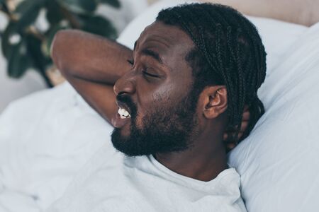 Unhappy African American man suffering from neck pain while lying in bedの写真素材