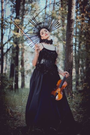 Low angle view of young woman in witch costume standing on forest background, holding violin