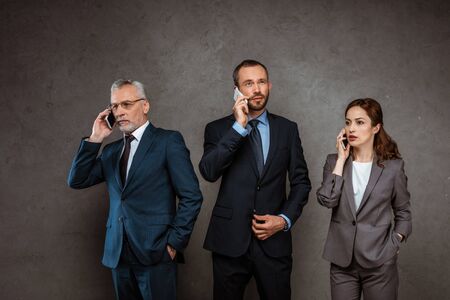 Handsome businessmen and attractive businesswoman in formal wear talking on smartphones on grey...