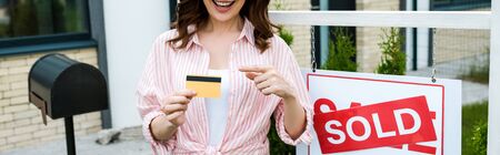 Panoramic shot of cheerful woman pointing with finger at credit card near board with sold letters
