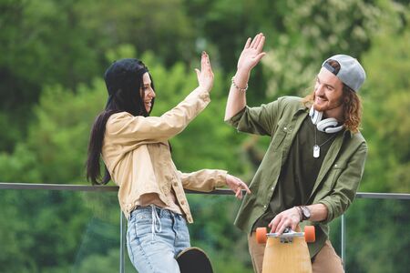 Happy man with skateboard high five to brunette woman, standing on green trees background
