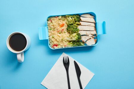 Top view of cup of coffee, disposable fork with knife and lunch box with risotto, chicken and broccoli on blue backgroundの写真素材