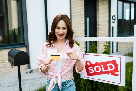 Cheerful woman pointing with finger at credit card near board with sold lettersの写真素材