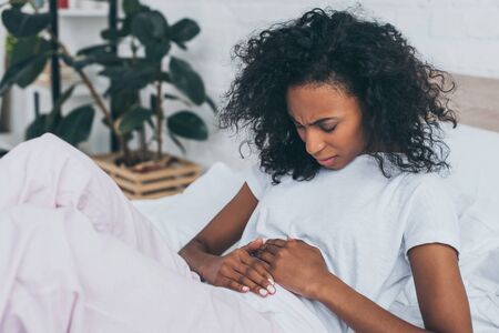 Unhappy African American woman suffering from abdominal pain while lying in bedの写真素材