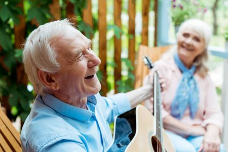 Selective focus of happy retired husband playing acoustic guitar near senior wifeの写真素材