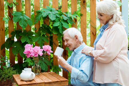 Cheerful retired woman standing near happy senior man looking at cup
