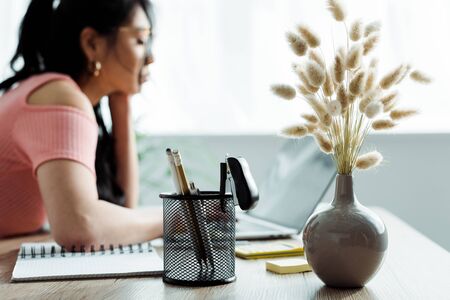 Selective focus of vase with willow branches and pen holder near Asian woman