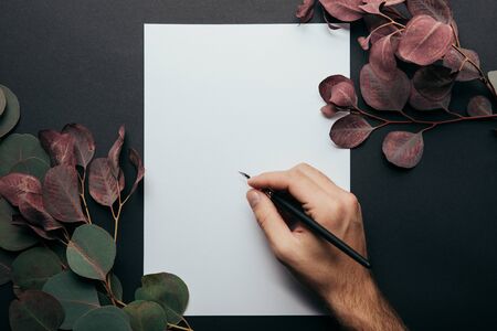Cropped view of man writing with fountain pen on paper on black background with eucalyptus branches