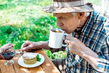 Senior man in straw hat drinking tea near sweet and tasty pancakesの写真素材