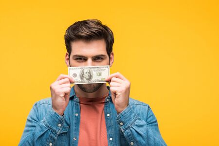 Handsome man covering face with money isolated on yellow background