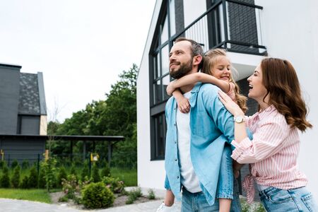 Happy man piggybacking daughter near cheerful wife and houseの写真素材