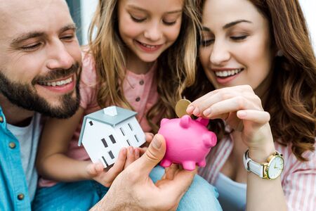 Selective focus of happy woman putting golden coin in piggy bank near husband and daughterの写真素材