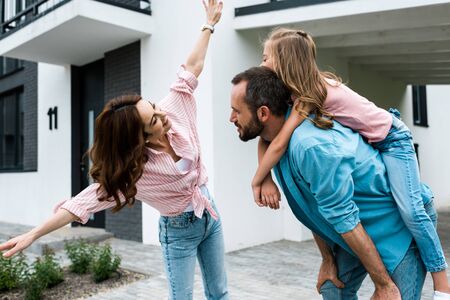 Happy man piggybacking daughter near cheerful wife with outstretched handsの写真素材