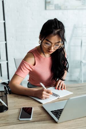 Attractive Asian woman in glasses writing in notebook near gadgetsの写真素材