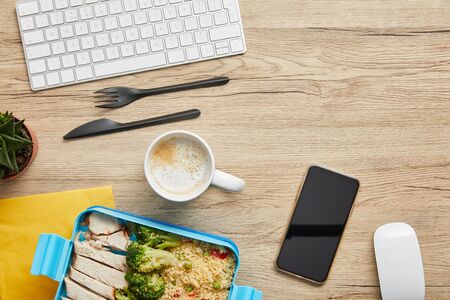 Top view of lunch box with tasty meal, hot beverage and digital devices on wooden table backgroundの写真素材