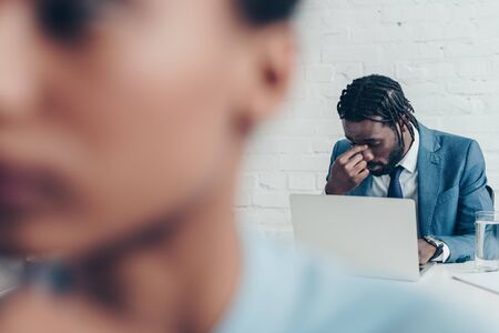 Selective focus of African American manager suffering from headache while sitting in office near colleagueの写真素材