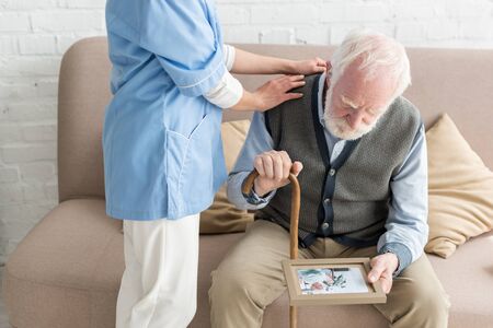 Nurse putting hands on upset and grey haired man with photo frameの写真素材