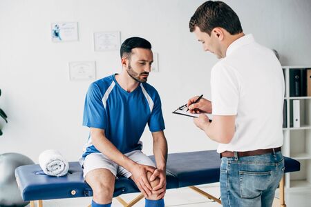 Physiotherapist writing diagnosis while football player sitting on Massage Table in hospitalの写真素材