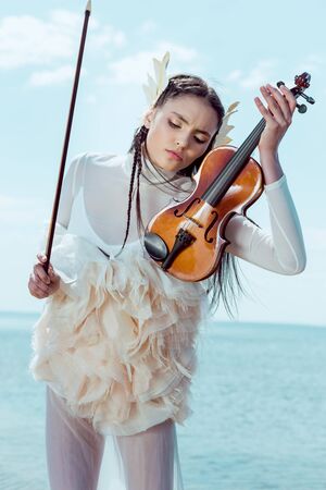 Tender woman in white swan costume standing on blue sky background with violin
