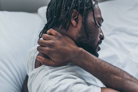 Young African American man with dreadlocks suffering from neck pain in bedroomの写真素材