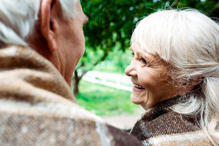 Selective focus of happy senior woman in plaid blanket looking at husbandの写真素材