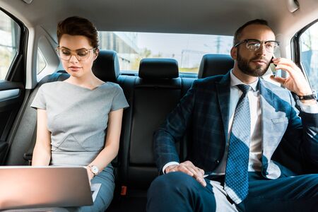 Attractive businesswoman using laptop near businesswoman talking on smartphone in car