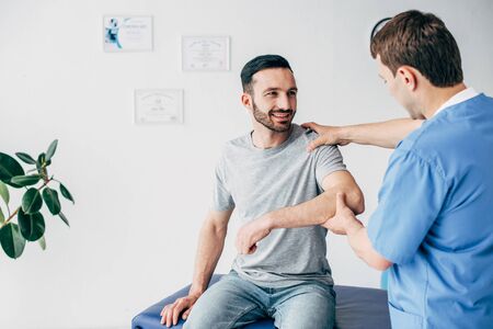 Smiling patient sitting on couch and doctor examining patient shoulder in massage cabinet at clinicの写真素材