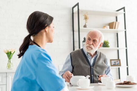 Calm grey haired man looking at nurse, sitting on kitchen at homeの写真素材