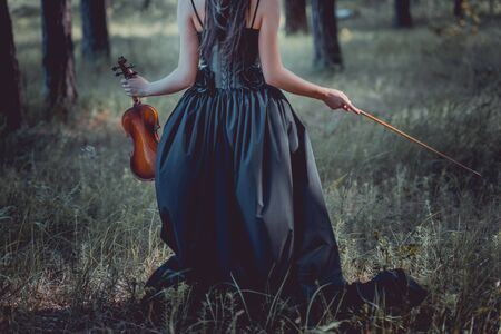 Back view of woman in witch costume walking on forest, holding violinの写真素材