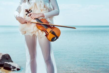 Cropped view of tender woman in white swan costume with violin standing on blue sky and river backgroundの写真素材
