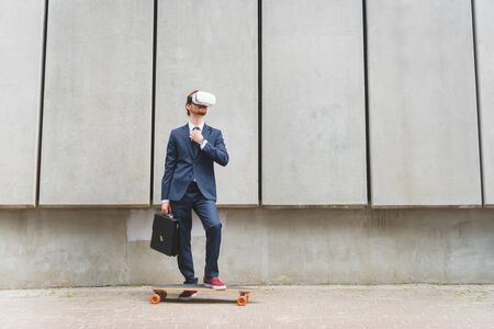 Businessman in formal wear and vr headset standing near longboard on concrete wall backgroundの写真素材