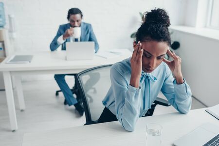 Selective focus of African American businesswoman suffering from headache while sitting in office near colleague drinking coffeeの写真素材