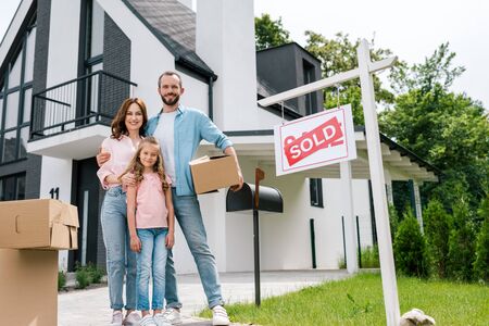 Happy man holding box and standing with wife and daughter near house and board with sold lettersの写真素材