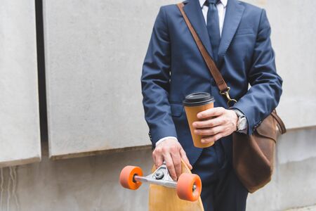 Cropped view of businessman in formal wear standing with skateboard and paper cup in handsの写真素材