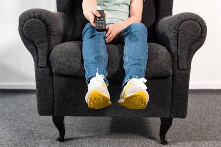 Boy in jeans sitting in armchair and holding remote controller
