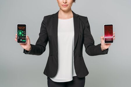 Partial view of mixed race businesswoman holding smartphones with trading courses and marketing analysis apps on screen isolated on grey backgroundの写真素材