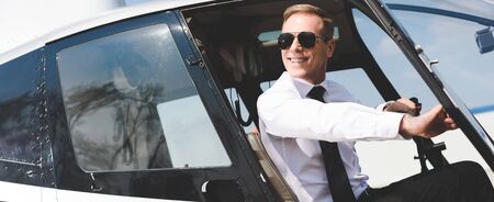 Panoramic shot of good-looking smiling Pilot in formal wear and sunglasses in helicopter cabin