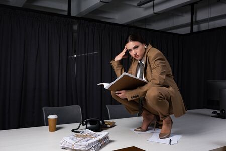 Stylish businesswoman in suit sitting on table, holding documents and looking at cameraの写真素材