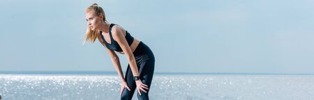 Panoramic shot of attractive and tired sportive woman standing on beach near sea