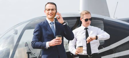 Panoramic shot of businessman with coffee to go talking on smartphone while pilot looking at watch...