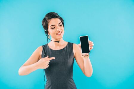 Smiling mixed race call center operator pointing wit finger at smartphone with blank screen isolated...