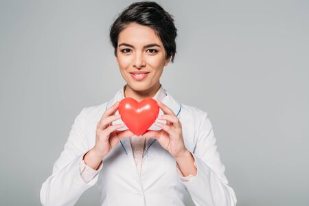 Cheerful mixed race doctor holding heart model while smiling at camera isolated on grey backgroundの写真素材