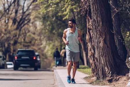 Handsome sportsman running along pavement near roadway and listening music in earphones.の写真素材