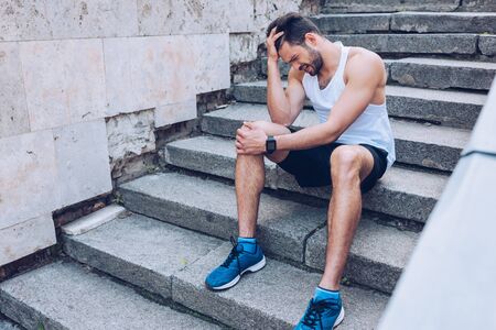 Injured sportsman suffering from pain while sitting on stairs and holding hand near headの写真素材