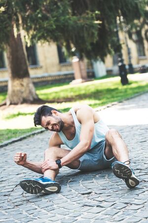 Young sportsman suffering from pain while sitting on pavement and touching injured leg
