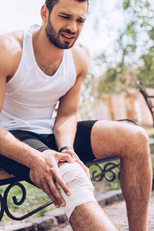 Upset sportsman sitting on bench and touching elastic bandage on injured kneeの写真素材