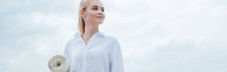 Panoramic shot of cheerful young blonde woman standing near sea and holding yoga matの写真素材