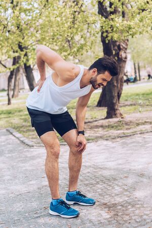 Young man in sportswear and sneakers suffering from hurt while touching injured back