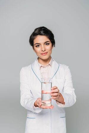 Attractive mixed race doctor holding glass of water and looking at camera isolated on grey backgroundの写真素材